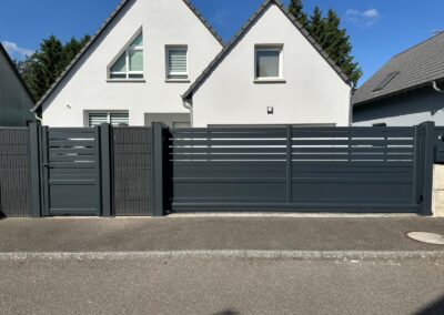 Portail et portillon aluminium anthracite à lames ajourées, devant une maison blanche à deux pignons